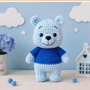 Könnte beinhalten: Ein hellblau gehäkelter Bär mit einem dunkelblauen Pullover, schwarzen Kunststoffaugen und einer schwarzen gestickten Nase. Der Bär hat abgerundete Ohren, eine strukturierte Garnoberfläche und einen neutralen Gesichtsausdruck.