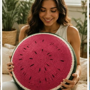Könnte beinhalten: Ein rundes, gehäkeltes Kissen, das wie eine Wassermelonenscheibe gestaltet ist. Die Mitte ist leuchtend rosa mit verstreuten schwarzen Garnstichen, die Samen darstellen, umgeben von einem dünnen weißen Ring und einem dunkelgrünen Rand.