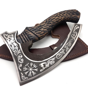 May include: A hand axe with a dark, textured wooden handle and a steel blade featuring intricate silver-toned scrollwork engravings and a central Vegvisir symbol. The axe rests on a brown leather sheath with visible stitching and a metal snap closure.