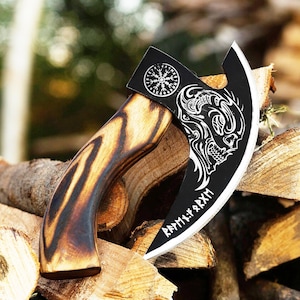 May include: A hand axe with a curved wooden handle featuring dark grain patterns and a black metal blade. The blade displays a white graphic of a skull, a circular Vegvisir symbol, and runic inscriptions along the sharpened edge.