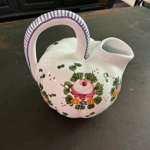 May include: A white ceramic pitcher with a rounded, pumpkin-like shape and a curved handle featuring blue and white stripes. The body is decorated with hand-painted floral motifs in shades of pink, green, orange, and yellow, with a wide spout.
