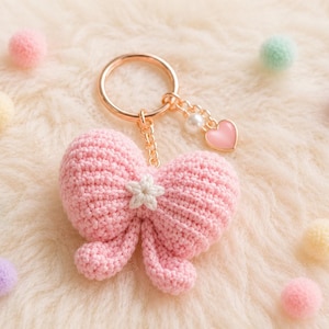 May include: A light pink crocheted bow keychain featuring a small white flower at the center. The keychain includes a gold-toned metal ring, a short chain, a single white pearl bead, and a small pink heart-shaped charm attached to the ring.