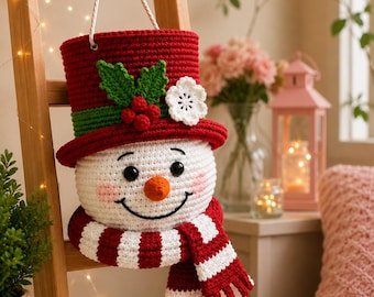 May include: A crocheted snowman wall hanging featuring a white head with black eyes, a carrot-colored nose, and pink cheeks. It wears a red top hat with a green band, green holly leaves, red berries, and a white flower, plus a red and white striped scarf.