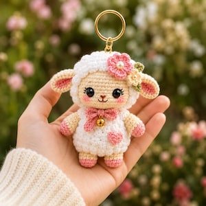 May include: A white crocheted lamb keyring featuring a pink bow, a small gold bell, and a pink heart on its chest. The lamb has large black eyes, pink blush on its cheeks, and a small flower accessory on its head with a gold metal ring attached.