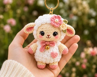 May include: A white crocheted lamb keychain featuring a pink bow, a small gold bell, and a pink heart on its chest. The lamb has large black eyes, pink blush on its cheeks, and a small flower accessory on its head with a gold metal ring attached.