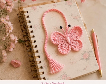 May include: A light pink crocheted butterfly bookmark with a long braided tail ending in a tassel. The butterfly features two black bead antennae and is resting on a spiral-bound notebook with a floral pattern, accompanied by a matching pink pen.