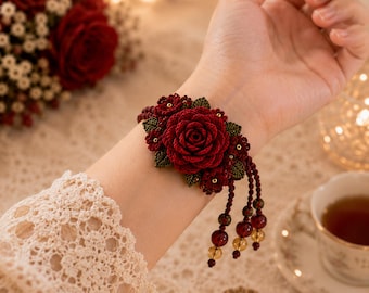 May include: A dark red crochet rose bracelet featuring intricate floral detailing with small green leaves and three dangling beaded strands. The bracelet is crafted from fine thread and includes small dark red and amber-colored beads at the ends.