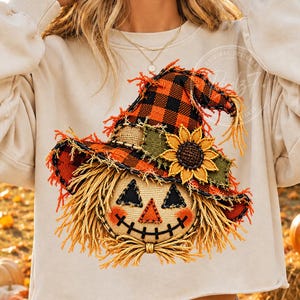 May include: A beige long-sleeve sweatshirt featuring a graphic of a scarecrow face wearing a plaid orange and black witch hat. The scarecrow has a burlap-textured face with triangular eyes, a triangular nose, a stitched mouth, and straw hair.