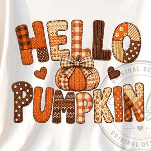 以下が含まれることがあります： クリーム色の半袖Tシャツ。オレンジ、ブラウン、ベージュの様々なパッチワーク風パターンで「HELLO PUMPKIN」の文字が描かれています。中央にはチェック柄のリボンが付いたカボチャのグラフィックがあり、その両脇に小さなハートが配置されています。