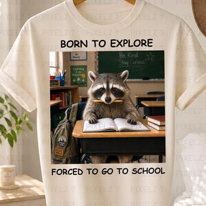 May include: A cream-colored t-shirt featuring a graphic of a raccoon sitting at a school desk with an open book and a pencil in its mouth. The text above the image reads BORN TO EXPLORE and the text below the image reads FORCED TO GO TO SCHOOL.