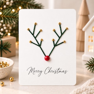 May include: A white rectangular greeting card featuring a reindeer antler design created with dark green thread and small gold beads at the tips. A single red bead serves as the nose. The text Merry Christmas is printed in black script below.