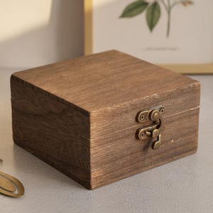 Pode incluir: Uma caixa quadrada de madeira com acabamento em grão de madeira castanho escuro e textura visível. A caixa possui um fecho de metal cor de latão na parte frontal. A tampa é plana e a caixa tem uma construção simples com padrões de grão.