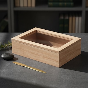 May include: A rectangular light-coloured wooden box featuring a clear glass lid. The box has a smooth, natural wood grain finish and a hinged top. Beside the box lies a single smooth dark grey stone and a slender gold-toned metal letter opener.