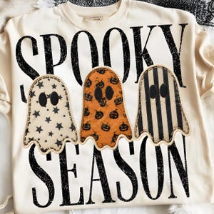 May include: A cream-colored sweatshirt featuring the black distressed text SPOOKY SEASON. Three ghost-shaped patches are centered on the front: one with black stars, one orange with black pumpkins, and one with black vertical stripes, all with black eyes.