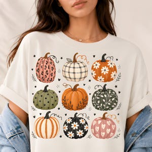 May include: A cream-colored short-sleeve t-shirt featuring a graphic print of nine pumpkins in a grid. The pumpkins display various patterns including leopard print, plaid, white daisies, black polka dots, orange leaves, white vines, stripes, and hearts.