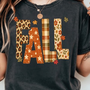 Op de afbeelding: Een antracietgrijs t-shirt met het woord FALL in grote blokletters. Elke letter heeft een eigen patroon: luipaardprint, oranje met witte sterren, groen en beige ruitpatroon, en een bloemendesign met oranje bloemen en bladeren.