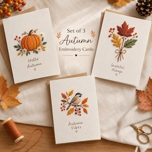 May include: A set of three off-white greeting cards featuring embroidered autumn designs. One card shows a pumpkin with flowers and the text Hello Autumn. Another displays maple leaves with the text Grateful Always. The third shows a bird on a branch with the text Autumn Vibes.