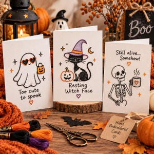May include: Three white greeting cards featuring hand embroidery. The first shows a ghost with heart sunglasses and a pumpkin bucket with the text Too cute to spook. The second shows a cat in a witch hat with a pumpkin and the text Resting Witch Face. The third shows a skeleton holding a coffee mug with the text Still alive... Somehow.