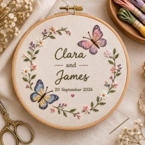May include: A circular wooden embroidery hoop containing off-white fabric with hand-stitched floral vines, two butterflies in pink and blue, and the text Clara and James 20 September 2026 with a small pink heart centered below the date.