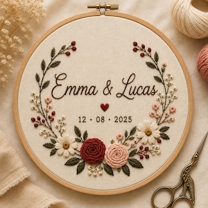 May include: A circular wooden embroidery hoop containing cream-colored fabric with hand-stitched floral wreath embroidery. The design features the names Emma & Lucas, a small red heart, and the date 12 . 08 . 2025 in dark brown thread.