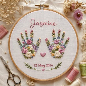 May include: A circular wooden embroidery hoop containing white fabric with hand-stitched floral designs shaped like two open hands. The text Jasmine is stitched in pink above the hands, with 12 May 2024 and small hearts stitched below.