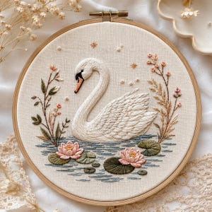 Può includere: Un telaio da ricamo circolare in legno contenente un pannello in tessuto con un cigno bianco ricamato a mano che galleggia su acqua blu. La scena presenta ninfee rosa, foglie verdi e delicati rametti floreali su entrambi i lati, con stelle.