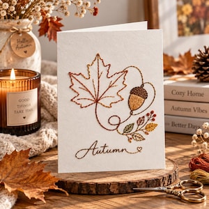 Puede incluir: Una tarjeta de felicitación blanca con un diseño bordado de una hoja de arce grande, una bellota y follaje pequeño. La palabra Autumn está bordada en letra cursiva en la parte inferior con un pequeño corazón. El bordado usa tonos marrones, naranjas y verdes.