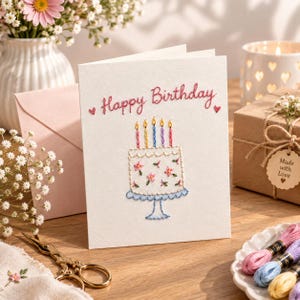 May include: A white greeting card featuring hand-embroidered text that reads Happy Birthday in pink thread. Below the text is an embroidered white cake with pink flowers, four colourful candles with yellow flames, and a light blue cake stand.