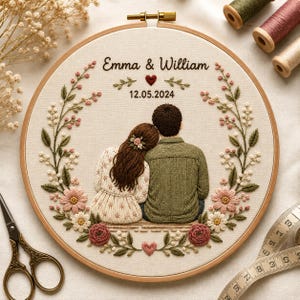 Op de afbeelding: Een ronde borduurring met een stoffen paneel met een handgestikte illustratie van een stel dat samen zit. De tekst "Emma & William" en "12.05.2024" staat erboven, omlijst door een bloemenkrans van roze en witte bloemen.