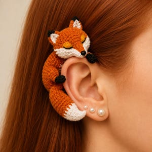 May include: A crochet fox ear cuff featuring a small orange and white fox with black-tipped ears and paws. The fox is designed to wrap around the outer edge of the ear, with its tail curving downward along the side of the earlobe.