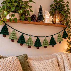 Puede incluir: Una guirnalda de ganchillo con pinos de color verde oscuro y verde claro alternados con troncos marrones, ensartados en un cordón de lana verde oscuro. Los árboles tienen un diseño texturizado y escalonado, formando nueve figuras individuales.