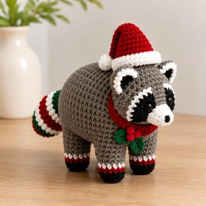 May include: A crocheted raccoon figurine featuring grey yarn, a black eye mask, and a white muzzle. It wears a red and white Santa hat, a red scarf with green holly leaves and red berries, and has a striped tail with red, white, and green colours.