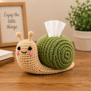 May include: A crochet snail tissue box cover featuring a beige head with black eyes, pink cheeks, and a smiling mouth. The shell is a large, spiralled green crochet piece with a white tissue protruding from the top centre of the snail's back.