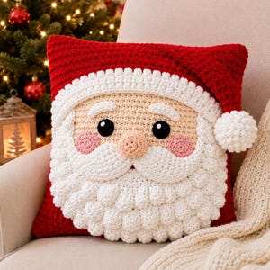 May include: A square crochet pillow featuring a Santa Claus face design. The pillow is primarily red with a white crocheted beard, mustache, eyebrows, and a hat brim. It includes black eyes, a peach-colored nose, and pink cheeks with a white pom-pom.