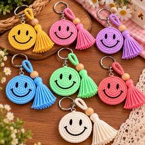 Pode incluir: Uma coleção de sete porta-chaves circulares de croché, cada um com uma cara sorridente bordada com olhos e boca pretos. Cada porta-chaves inclui uma borla a condizer, uma conta de madeira natural e um anel de metal prateado.