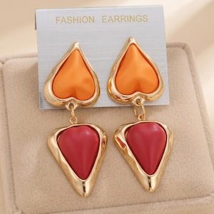 May include: A pair of drop earrings on a card labelled FASHION EARRINGS. Each earring features a gold-tone metal frame holding an orange heart-shaped top piece and a red heart-shaped bottom piece, connected by a small gold-tone metal jump ring.