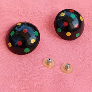 May include: A pair of round, black stud earrings featuring a glossy finish with hand-painted polka dots in red, yellow, and green. The set includes two clear plastic earring backs with metal posts for secure attachment to the earlobes.
