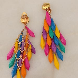 May include: A pair of gold-tone clip-on earrings featuring a cascade of multi-coloured, diamond-shaped plastic pendants in shades of pink, orange, yellow, blue, purple, and teal, suspended from a decorative gold-tone metal filigree structure.