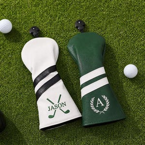 Könnte beinhalten: Zwei Golfschlägerhauben, eine weiße mit schwarzen Streifen und dem Namen JASON über gekreuzten Golfschlägern, und eine dunkelgrüne mit weißen Streifen und einem Buchstaben A in einem Lorbeerkranz, dazu zwei Golfbälle und zwei Schläger.