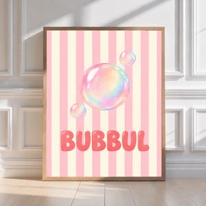 May include: A framed wall art print featuring a background of vertical light pink and cream stripes. Three iridescent, translucent bubbles float in the center. Below the bubbles, the word BUBBUL is printed in bold, rounded, coral-pink letters.