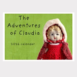 May include: A calendar with a green background featuring the text "The Adventures of Claudia" and "2026 calendar." A vintage doll with a red dress and a white-trimmed hood is on the right side of the calendar.