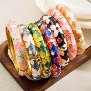 May include: A collection of eight hinged metal bangles arranged in a row, each featuring a vibrant enamel floral pattern. The bracelets display various color combinations including pink, grey, green, blue, black, and orange with white petals.