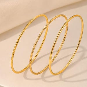 Puede incluir: Un conjunto de tres brazaletes finos de color dorado. El primer brazalete presenta un patrón texturizado de corte de diamante. El segundo brazalete tiene un diseño grabado repetitivo. El tercer brazalete es liso y pulido. Cada pulsera circular es de metal dorado.