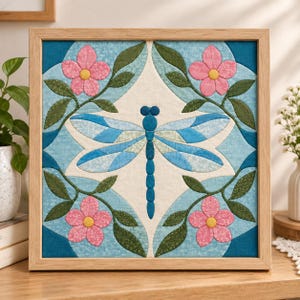 May include: A square fabric wall art piece in a light wood frame featuring a central blue dragonfly with segmented wings. The design includes four pink flowers with yellow centres and green leaves set against a cream and blue patterned background.