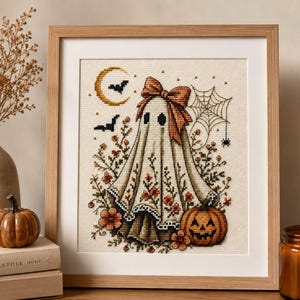 May include: A framed cross-stitch embroidery featuring a ghost wearing a large orange bow, surrounded by floral patterns, a jack-o'-lantern, a spider web with a spider, two bats, and a crescent moon on a light beige fabric background.