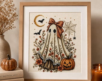 Può includere: Un ricamo a punto croce incorniciato raffigurante una figura di fantasma centrale che indossa un grande fiocco arancione. Il disegno include una mezzaluna, due pipistrelli, una ragnatela, una zucca e accenti floreali nei toni dell'arancione e del marrone.