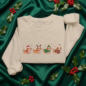 May include: A cream-colored crewneck sweatshirt featuring a graphic print of four brown dogs in a row. Each dog wears different holiday-themed attire, including a Santa hat, reindeer antlers, a Christmas tree costume, and a patterned sweater.