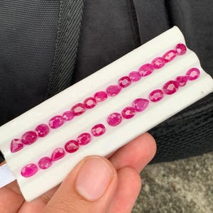 May include: A collection of small, faceted, vibrant pinkish-red rubies arranged in two rows on a white display card. The gemstones feature various shapes, including oval, round, and pear cuts, showcasing a brilliant, reflective surface.