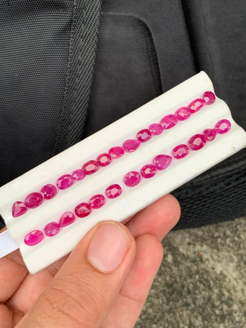 May include: A collection of small, faceted, vibrant pinkish-red rubies arranged in two rows on a white display card. The gemstones feature various shapes, including oval, round, and pear cuts, showcasing a brilliant, reflective surface.