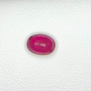 Peut inclure: Une pierre pr&eacute;cieuse taill&eacute;e en ovale, d'une couleur rose-rouge profonde et vibrante. La pierre pr&eacute;sente une surface &agrave; facettes qui refl&egrave;te la lumi&egrave;re, mettant en valeur sa qualit&eacute; translucide et ses bords arrondis sur un fond blanc textur&eacute;.
